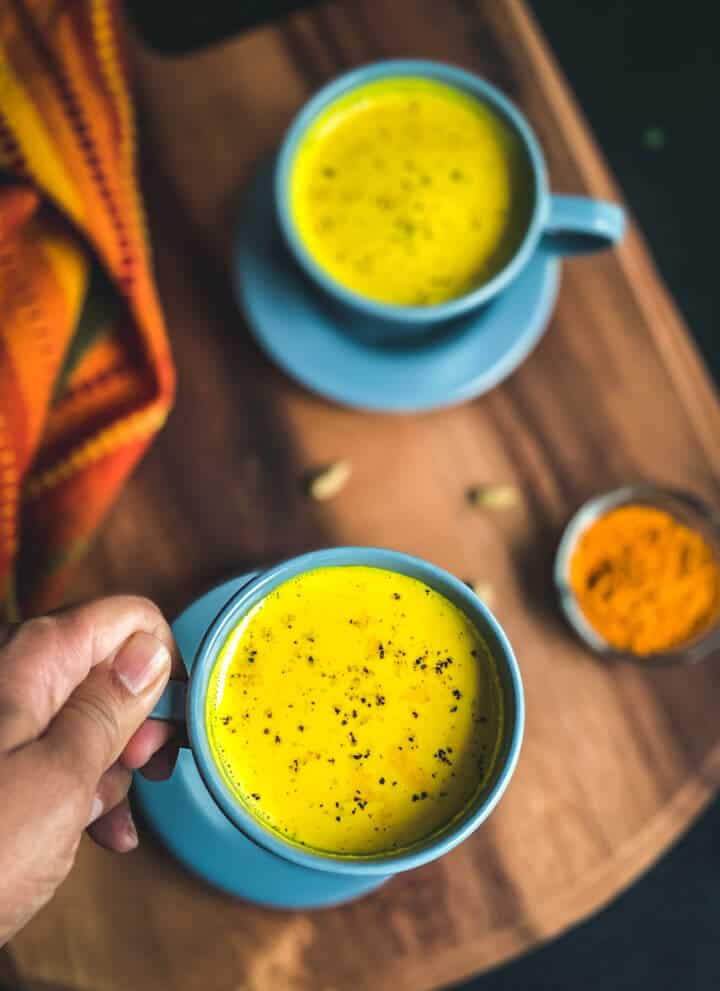 A hand holding a blue mug filled with haldi doodh over a blue saucer on a wooden counter next to a small bowl filled with turmeric.