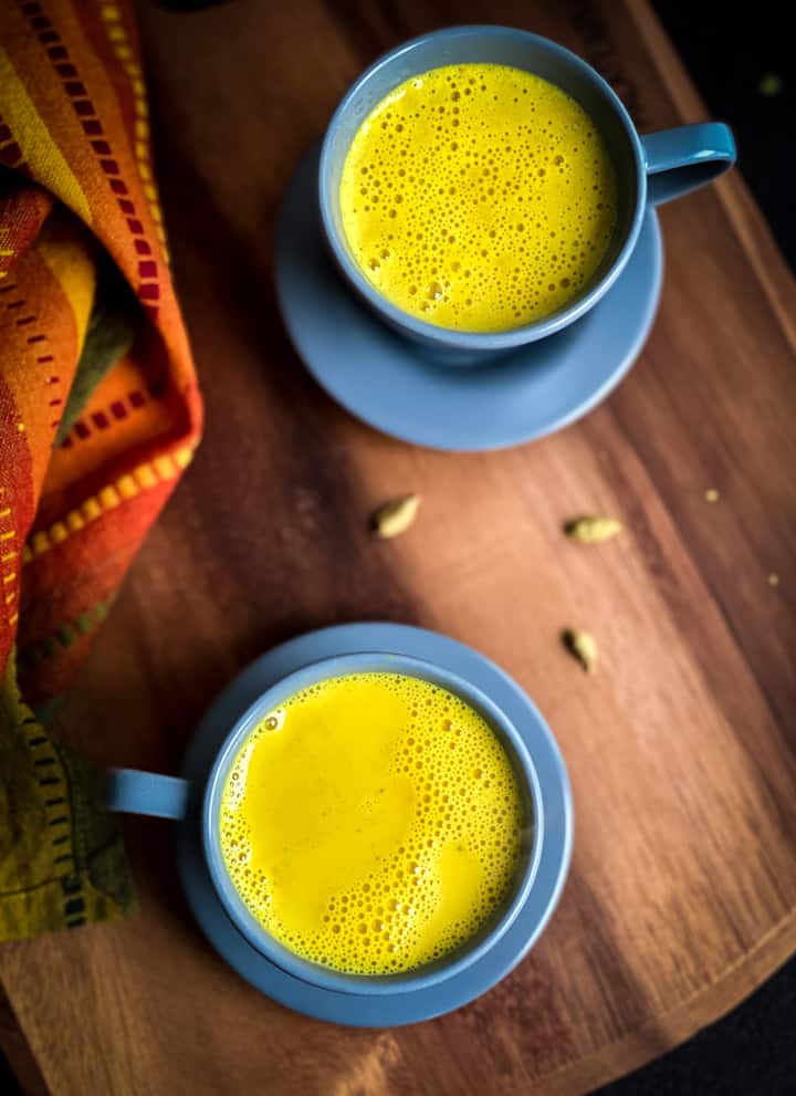 A wooden counter with two golden turmeric lattes in blue mugs.