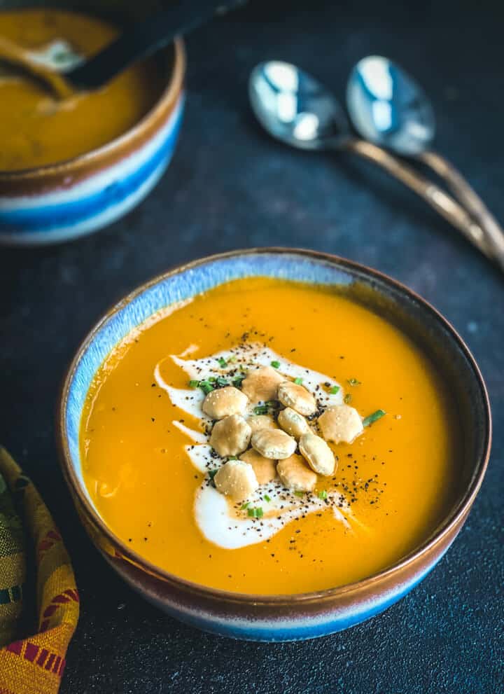 A blue bowl with a brown rim filled with vegan butternut squash soup and topped with coconut milk, chive, and oyster crackers on a blue counter with two spoons in the top right.