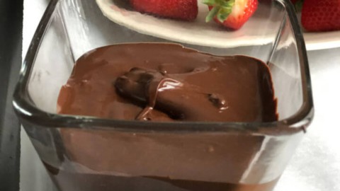 Melted chocolate in a tall glass with a plate of strawberries in the back.