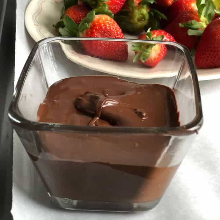 Melted chocolate in a tall glass with a plate of strawberries in the back.