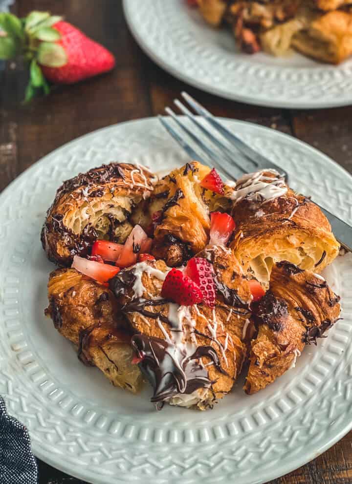 A white plate with a silver fork in the back and a large slice of chocolate croissant bread pudding topped with fresh fruit and drizzles of melted chocolate.