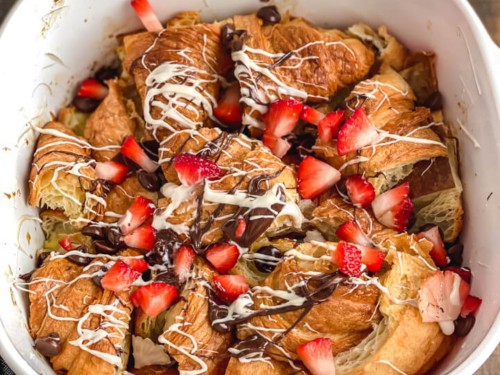 A white baking dish with chocolate croissant bread pudding topped with melted chocolate and fresh fruit.