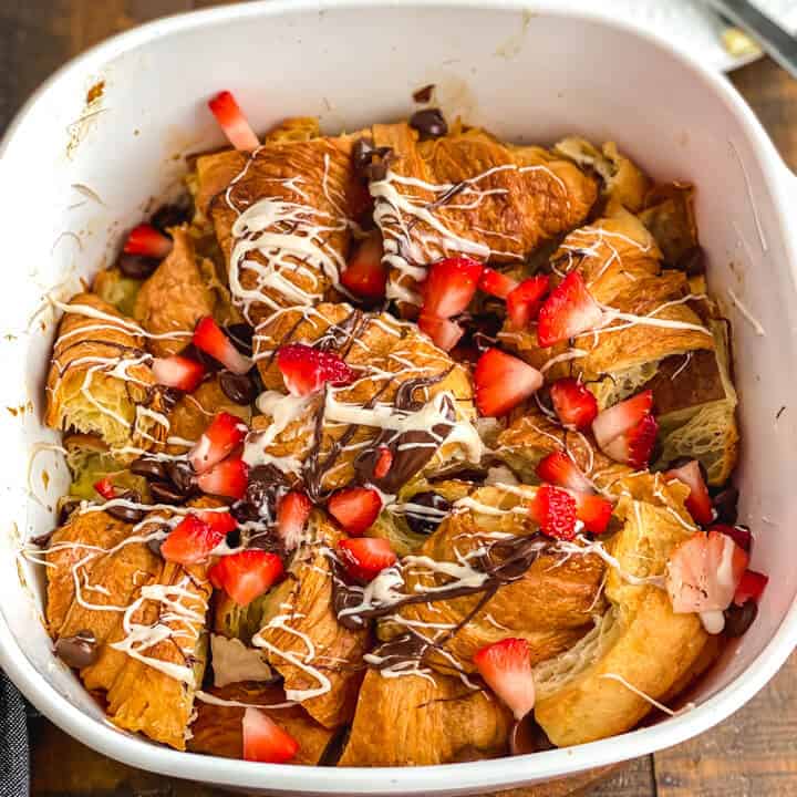 A square baking dish with croissant bread pudding drizzled with melted chocolate and topped with fresh fruit.