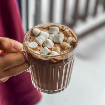 A square picture with a hand holding a clear glass mug of hot chocolate topped with marshmallows.