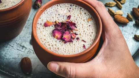 A hand holding a brown mug of thandai topped with rose petals over a silver platter with another mug of thandai to the left.