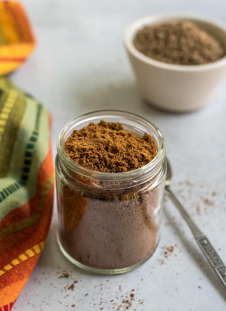 A glass jar with roasted cumin powder and a spoon to the right with cumin seeds in the back.