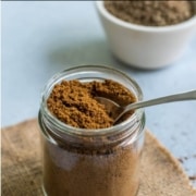 A glass jar with roasted cumin powder and a silver spoon and the words homemade roasted cumin powder at the top.