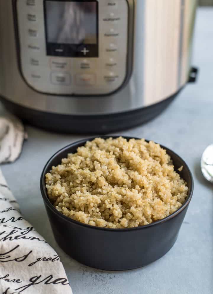 A black bowl filled to the top with fresh quinoa with a towel to the left of the bowl and an instant pot pressure cooker in the back left.