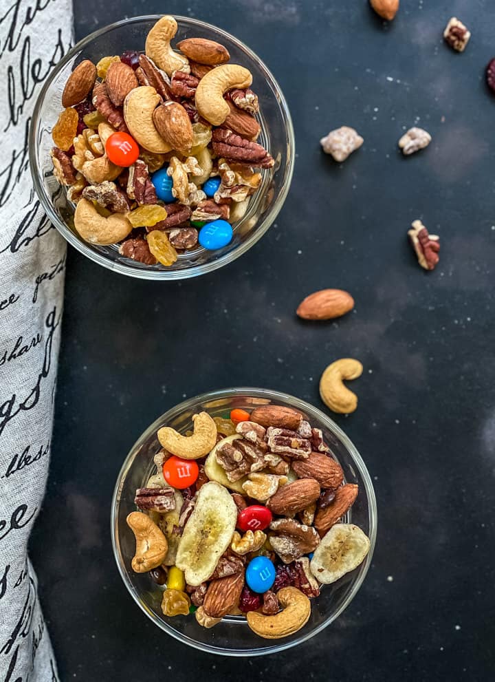 Two glass bowls of trail mix on a dark grey counter with other nuts sprinkled around.