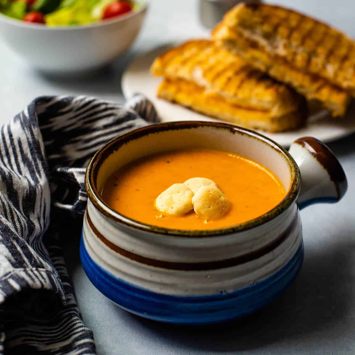 A small soup bowl with a handle on the right side filled with tomato basil soup and topped with oyster crackers with a grilled cheese sandwich in the back.