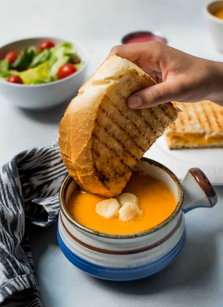 A small soup bowl with a handle on the right side filled with tomato basil soup and a hand holding a grilled cheese sandwich over the bowl of soup.