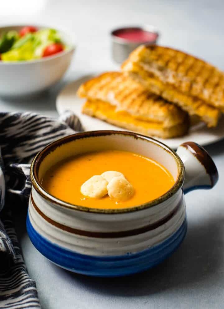 A small soup bowl with a handle on the right side filled with instant pot tomato basil soup and topped with oyster crackers with a side salad in the back left and a grilled cheese sandwich in the back right.