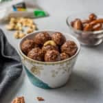 A small cup with chocolate date energy balls and a glass bowl of dates in the back right with a small pile of cashews in the back left.
