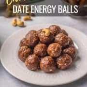 A white plate with a pile of date energy balls on top and a bowl of dates in the back right with a bag of cashews in the back left and the words Chocolate Date Energy Balls at the top.
