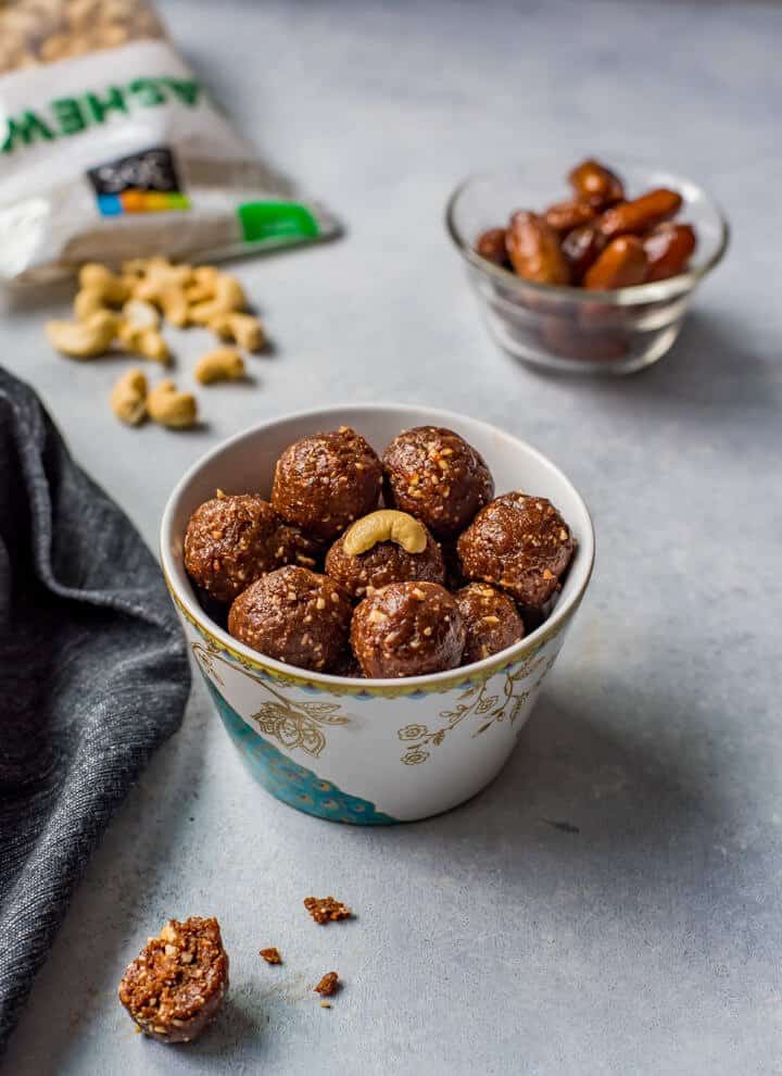 A small cup filled with date energy balls with dates in a small bowl in the back and cashews in the back left.