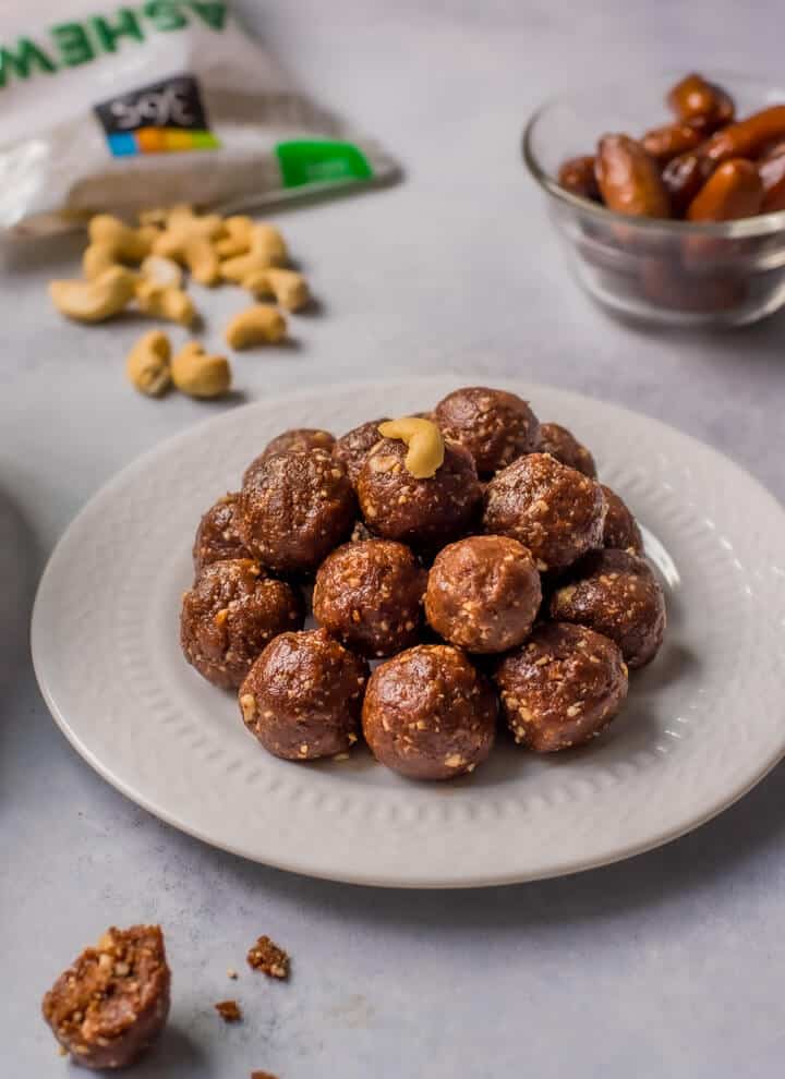 A white plate with a pile of date energy balls on top and a bowl of dates in the back right with a bag of cashews in the back left.