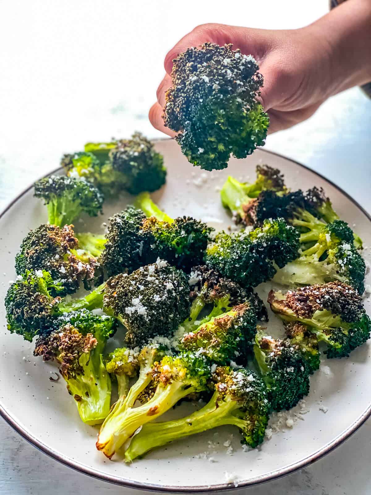 A hand shows a large floret of roasted broccoli, being pulled from a large white plate full of broccoli florets.