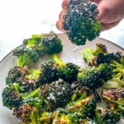 A hand picking up a piece of air fried broccoli from a white plate.