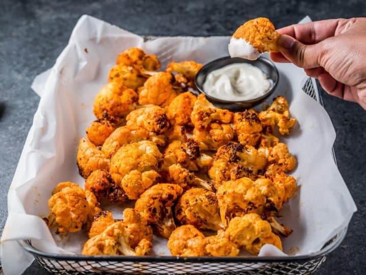 A hand dipping a piece of buffalo cauliflower into dipping sauce. A tray of cauliflower bites remains in the foreground.