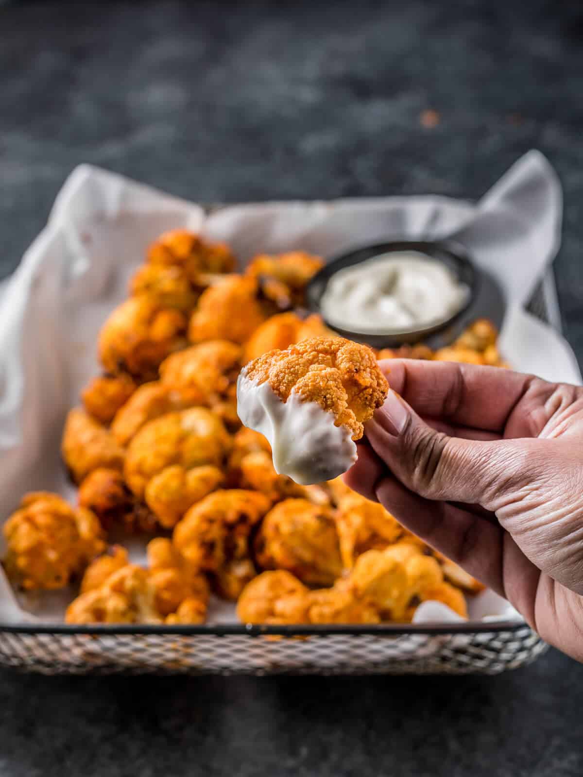 An air fryer tray filled with buffalo cauliflower in the background, with a hand dipping cauliflower into sauce.