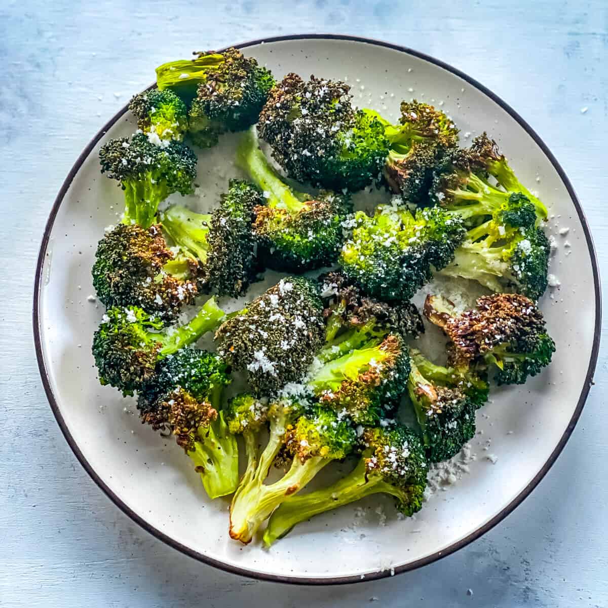 A white plate on a light blue background, holding a large amount of roasted broccoli florets.