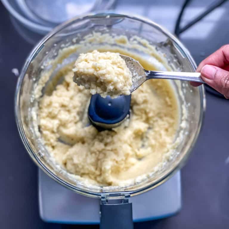 A food processor with garlic paste in it; a spoon lifts out a small portion.