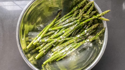 A silver bowl with trimmed asparagus topped with seasoning before tossing.
