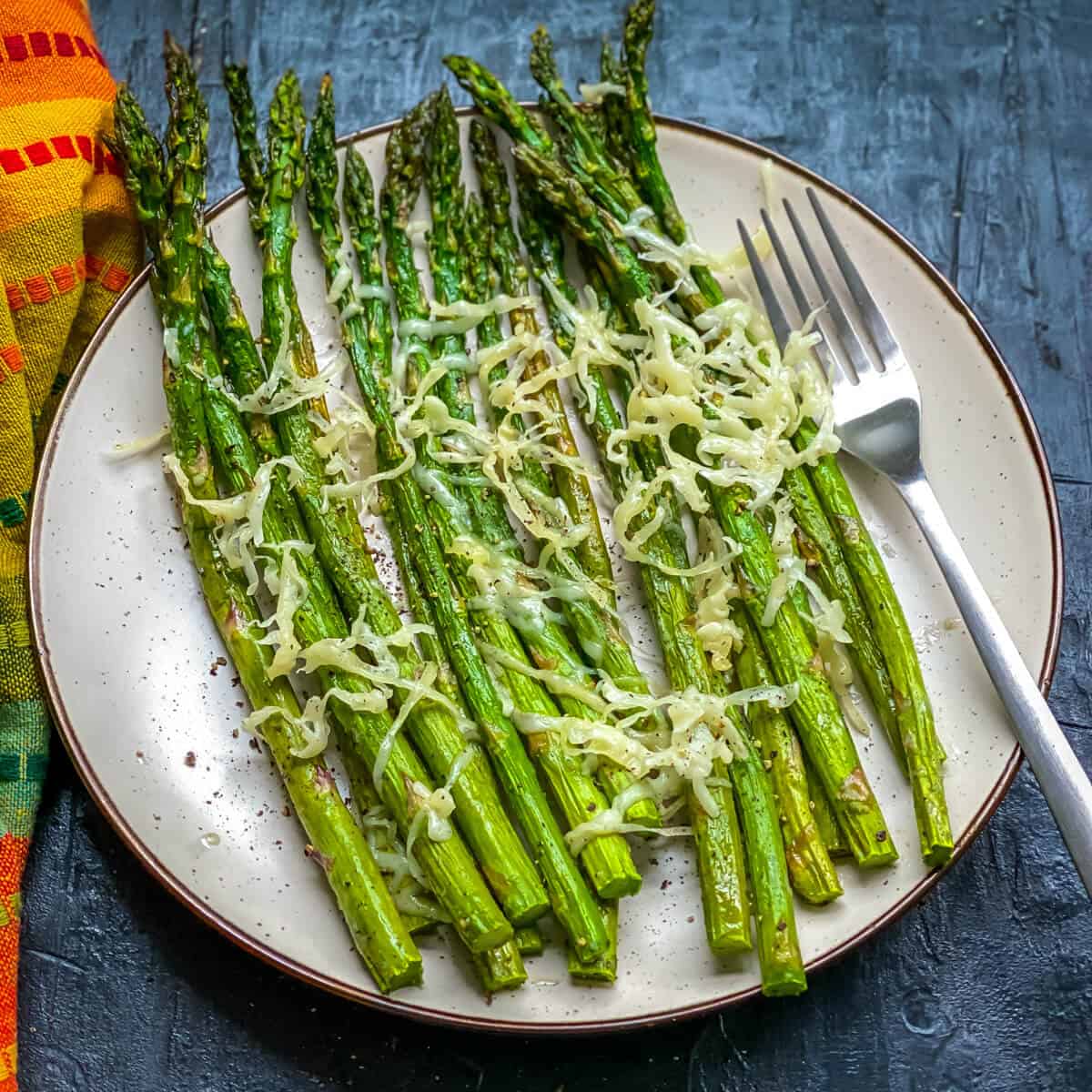 A cream plate on a blue counter with air fried asparagus topped with parmesan and a silver fork to the right.