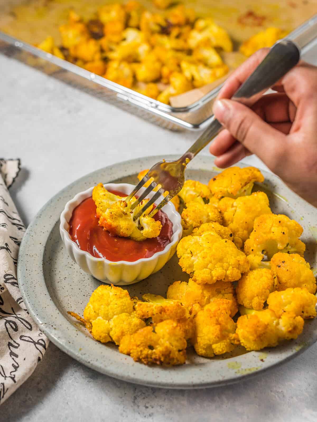 A fork grabbing a piece of cauliflower from a plate of roasted cauliflower, and dipping into a dish of ketchup.