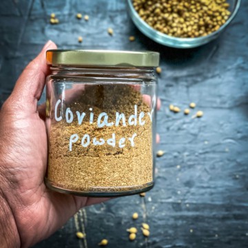 A hand holding a glass jar labeled coriander powder over a grey counter with coriander seeds.