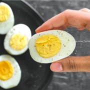 A hand holding a hard-boiled egg from the air fryer over a plate with other air fryer hard boiled eggs with the words How to Make Air Fryer Boiled Eggs at the top.