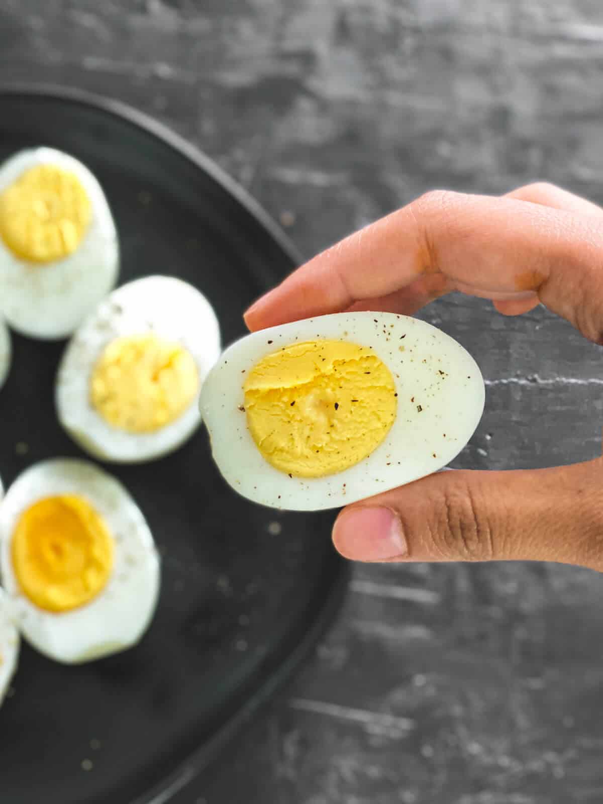A hand holding a hard-boiled egg from the air fryer over a plate with other air fryer hard boiled eggs