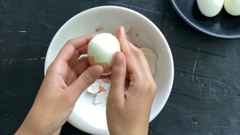 A pair of hands peeling the hard boiled egg over a white bowl.