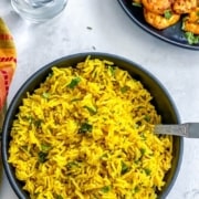 A black bowl of Indian Yellow Turmeric Rice with silver spoon on the right side of the bowl and a black plate of shrimp in the top right corner with the words How to Make Instant Pot Yellow Rice at the top.