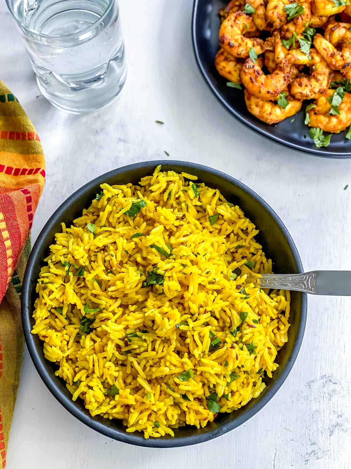 A black bowl of Indian Yellow Turmeric Rice with silver spoon on the right side of the bowl and a black plate of shrimp in the top right corner.