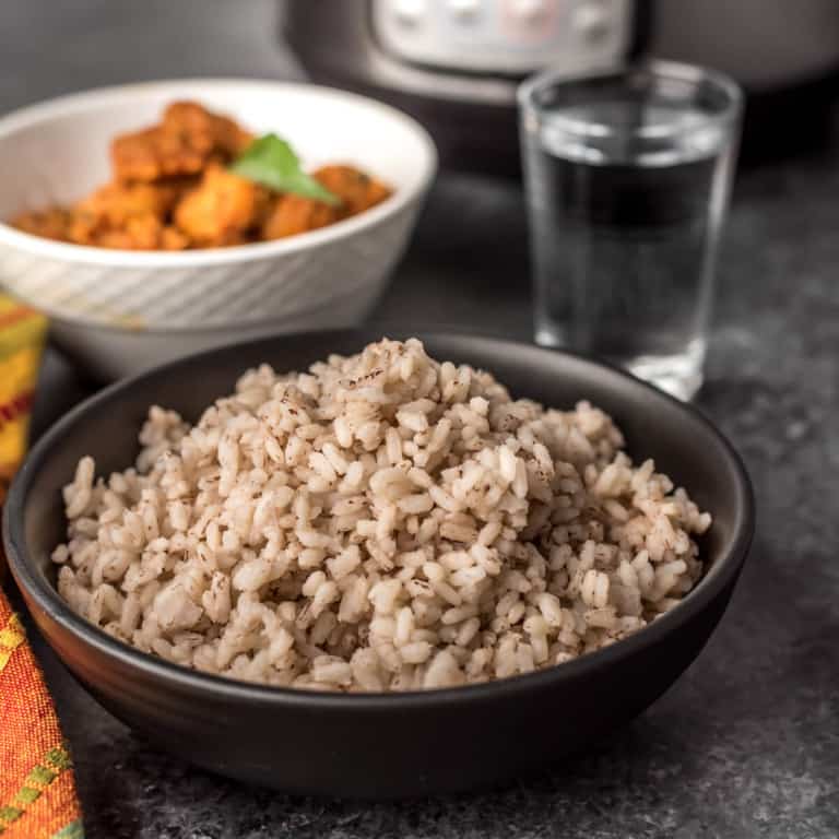 A black bowl with kreala matta rice after cooking in the Instant Pot, a cup of water in the back right, and a white bowl in the back left.