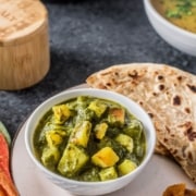 A white plate with a white bowl of aloo palak and a spoon in the front with fresh paratha in the back and the words Potato and Spinach Curry Aloo Palak in green at the top.
