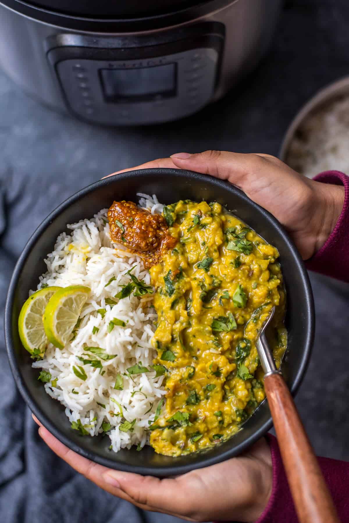 Two hands holding a black bowl with dal palak on the right side and fresh rice on the left side with a spoon on the right.