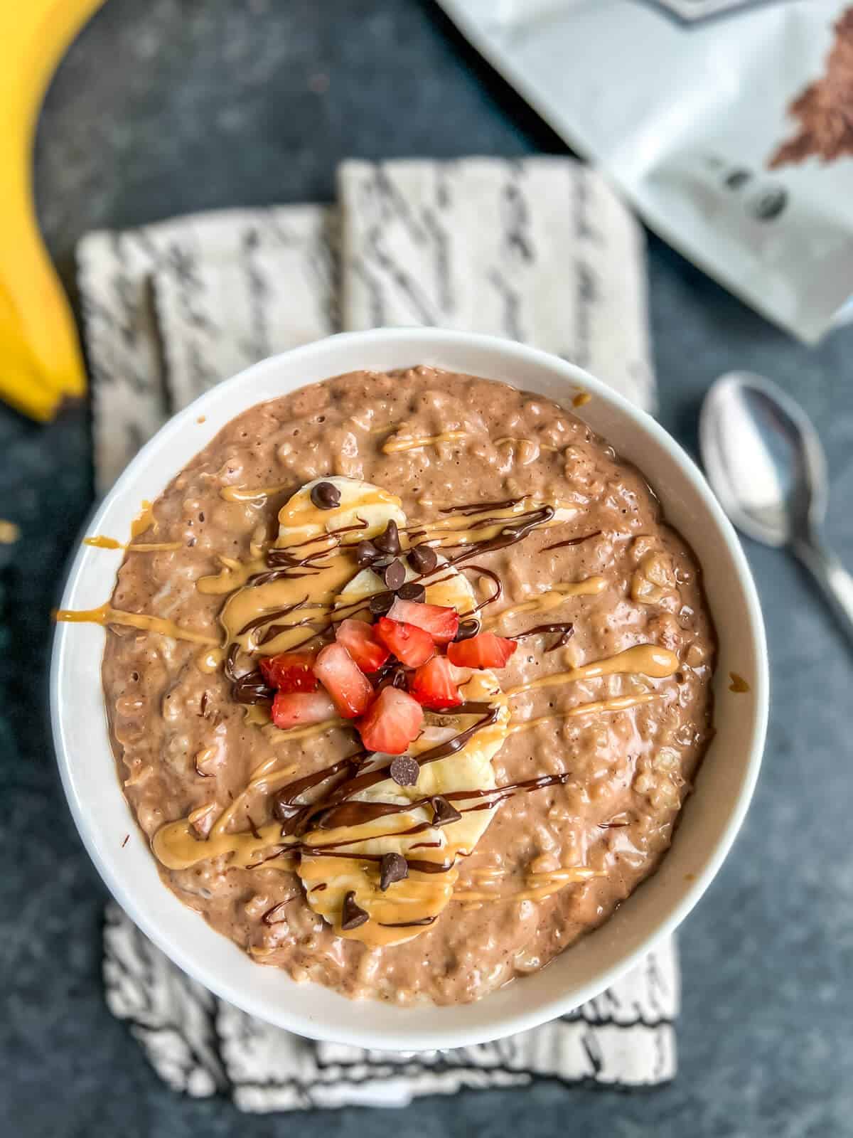 A white bowl with chocolate peanut butter oatmeal topped with slices of banana and some strawberry on a small white towel with a spoon to the right.