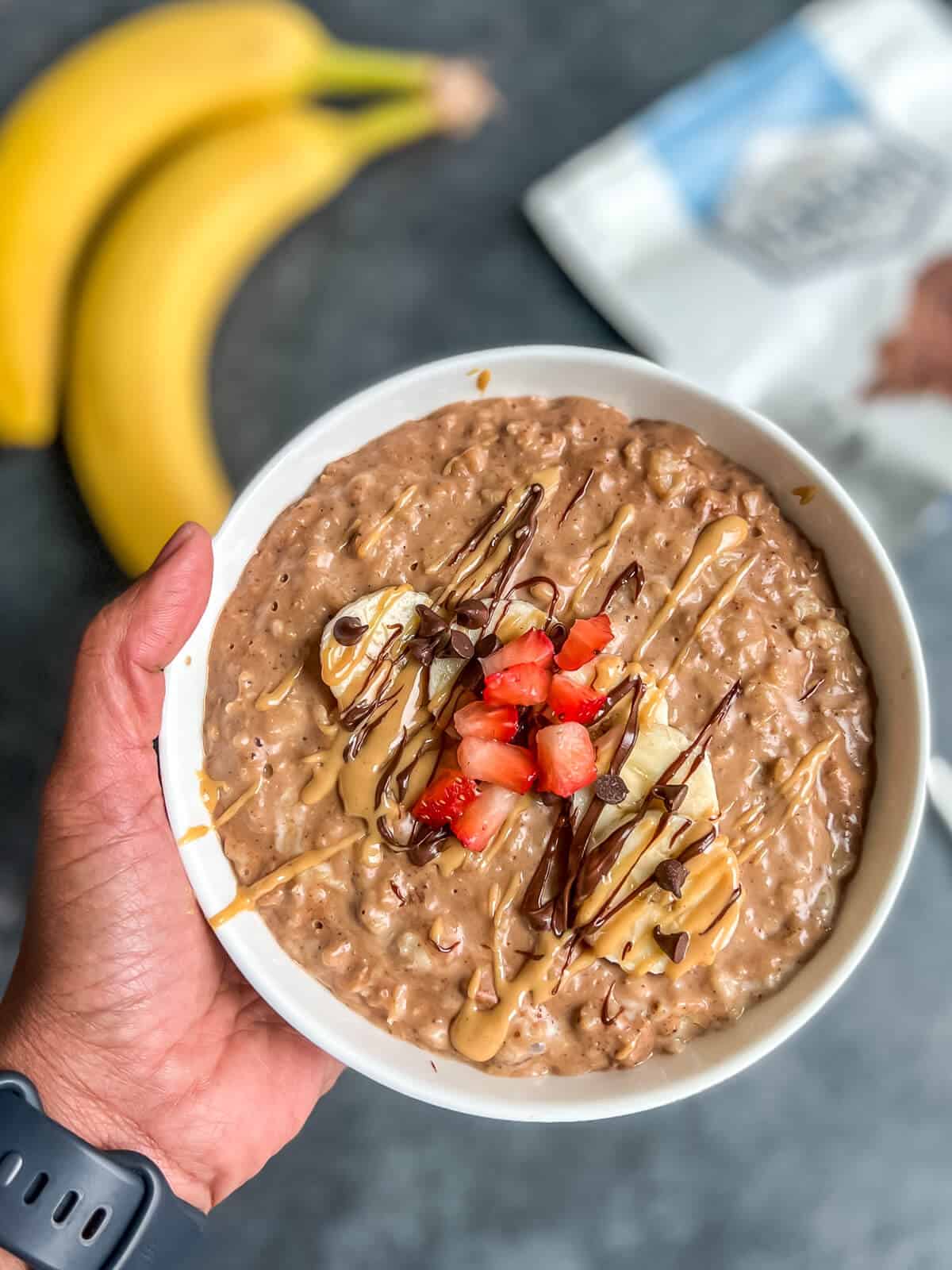 A hand holding a white bowl with chocolate peanut butter oats topped with banana and strawberry with a banana in the top left corner.