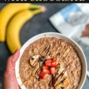 A hand holding a white bowl with chocolate peanut butter oats topped with banana and strawberry with a banana in the top left corner with the words Peanut Butter Oatmeal with Chocolate at the top.