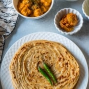 A white plate with a stack of lacha paratha topped with two green chilies in the front and a few white bowls in the back right corner and the words Laccha Paratha Crispy Whole Wheat Bread at the top.