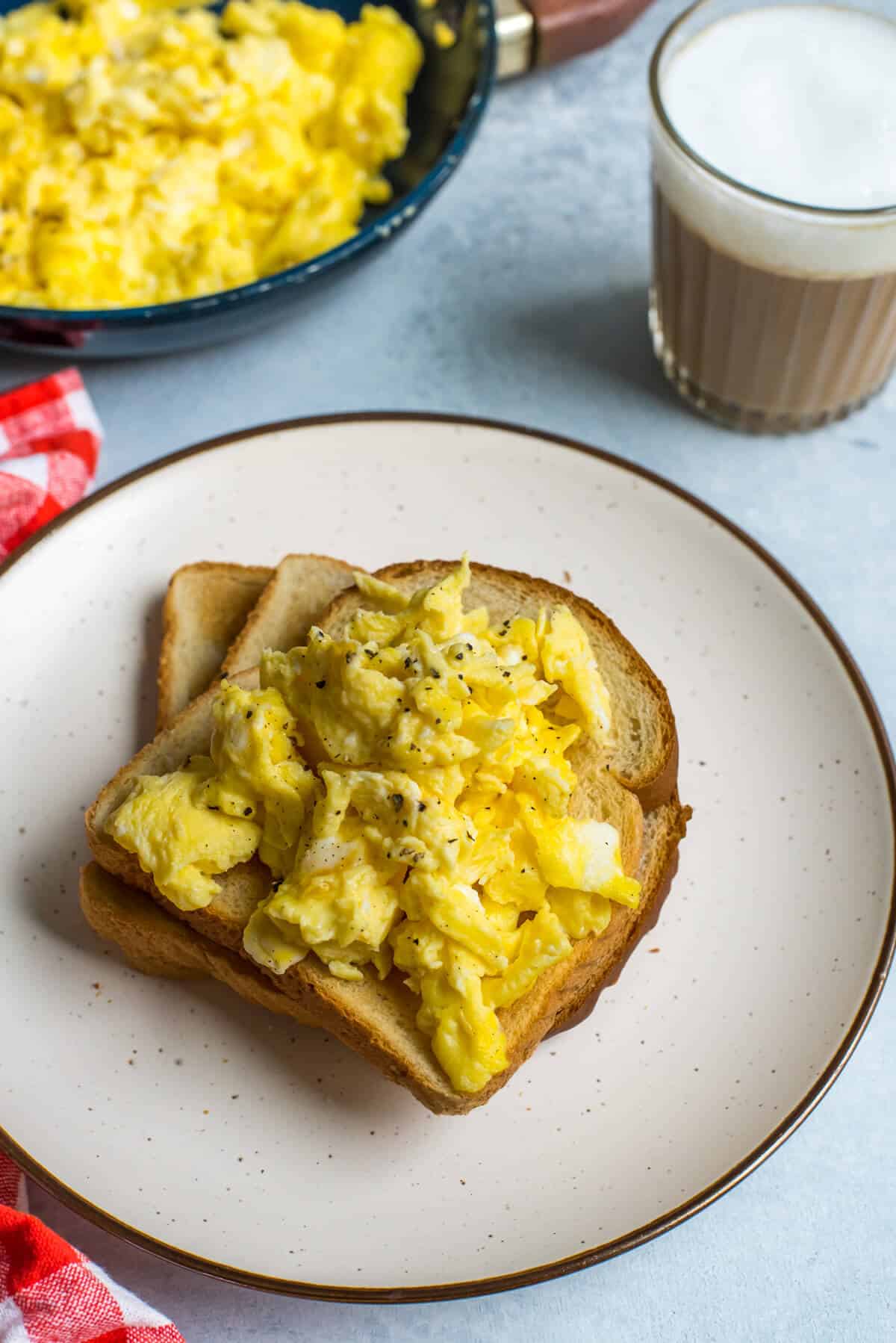 A white plate with a slice of toast topped with scrambled eggs and a cup of tea in the back right.