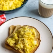 A white plate with a slice of toast topped with scrambled eggs and a cup of tea in the back right and the words Perfectly, Fluffy Scrambled Eggs at the top.