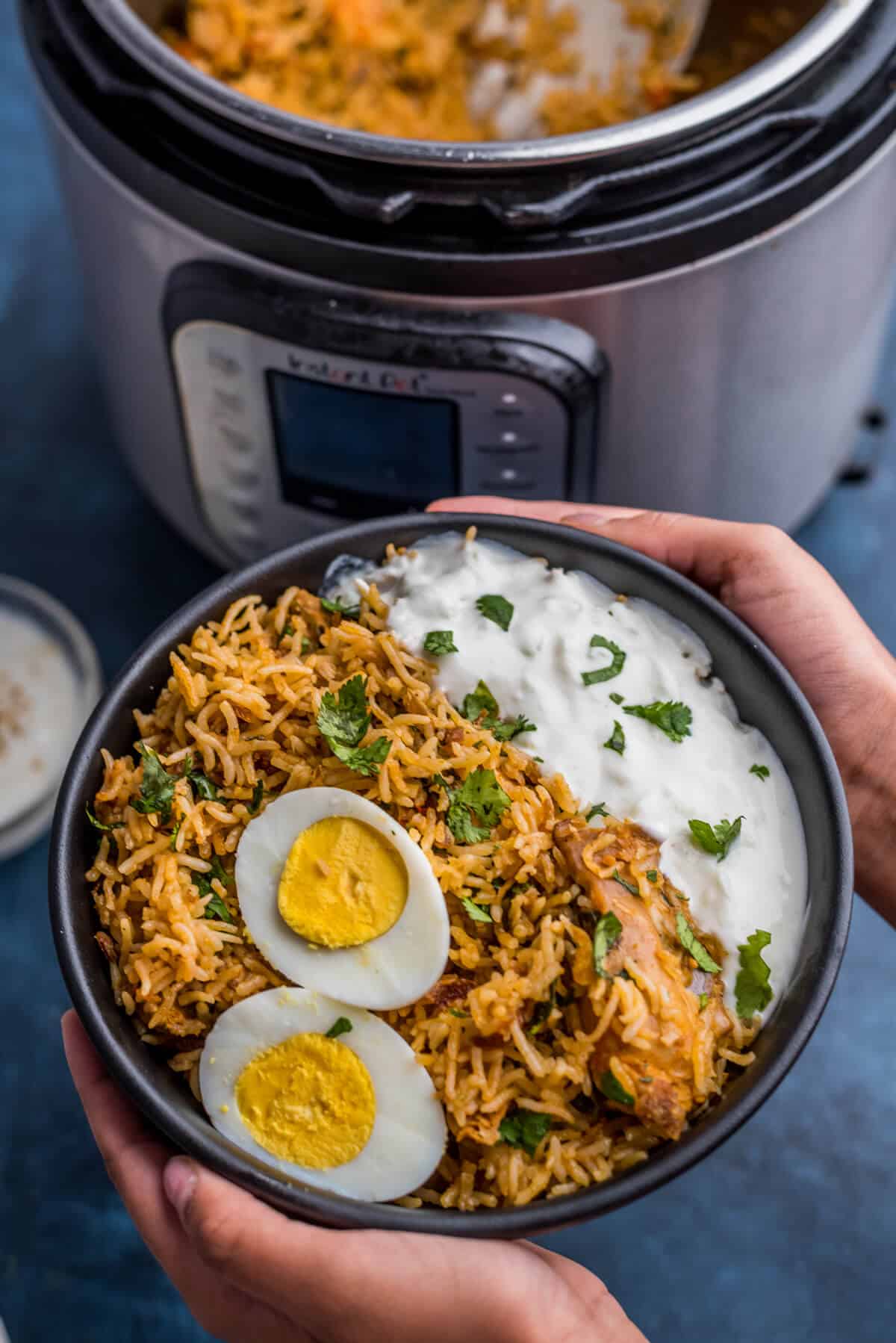 A black bowl with chicken biryani in the front and an Instant Pot in the back.