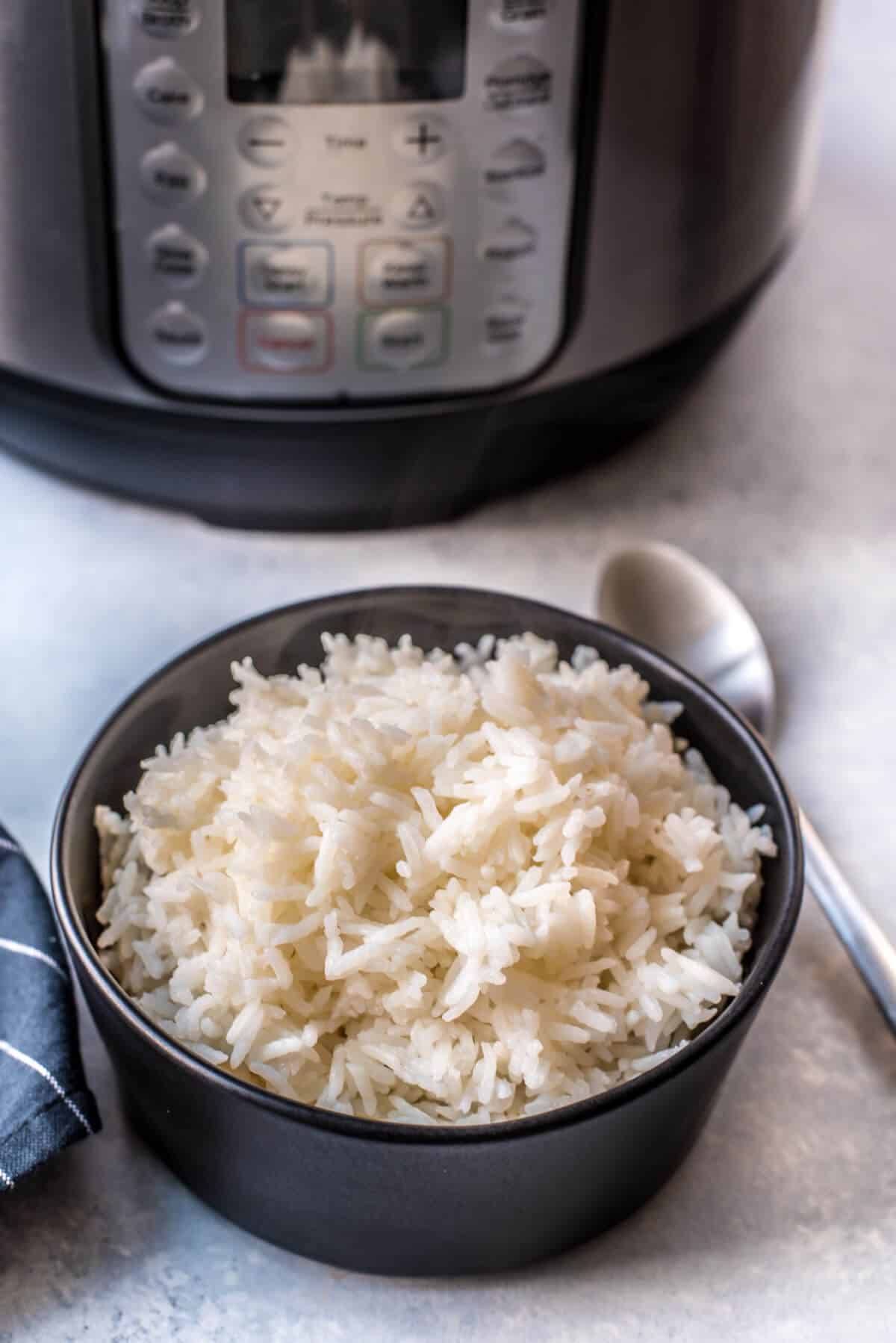 A black bowl with Instant Pot jasmine rice after cooking with a silver spoon to the right and an Instant pot pressure cooker in the back.