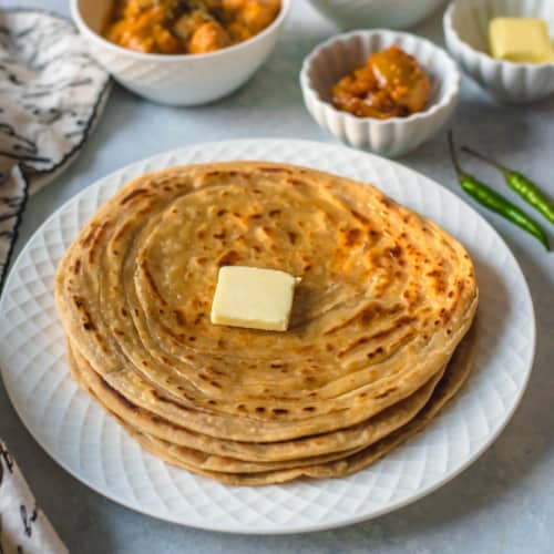 A white plate with a stack of lacha paratha topped with a pat of butter.