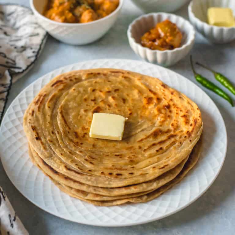 A white plate with a stack of lacha paratha topped with a pat of butter.