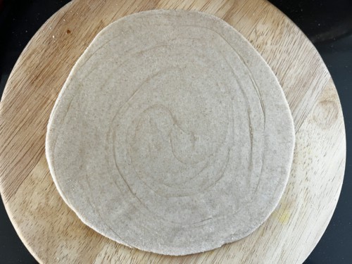 A round wooden counter with a rolled out disk of lacha paratha before being cooked in the pan.